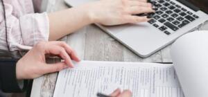 Close-up of hands typing on a laptop and reviewing business documents, focused on finance and legal tasks.
