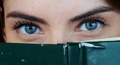 Close-up of a woman with blue eyes peeking over a book, portraying curiosity.